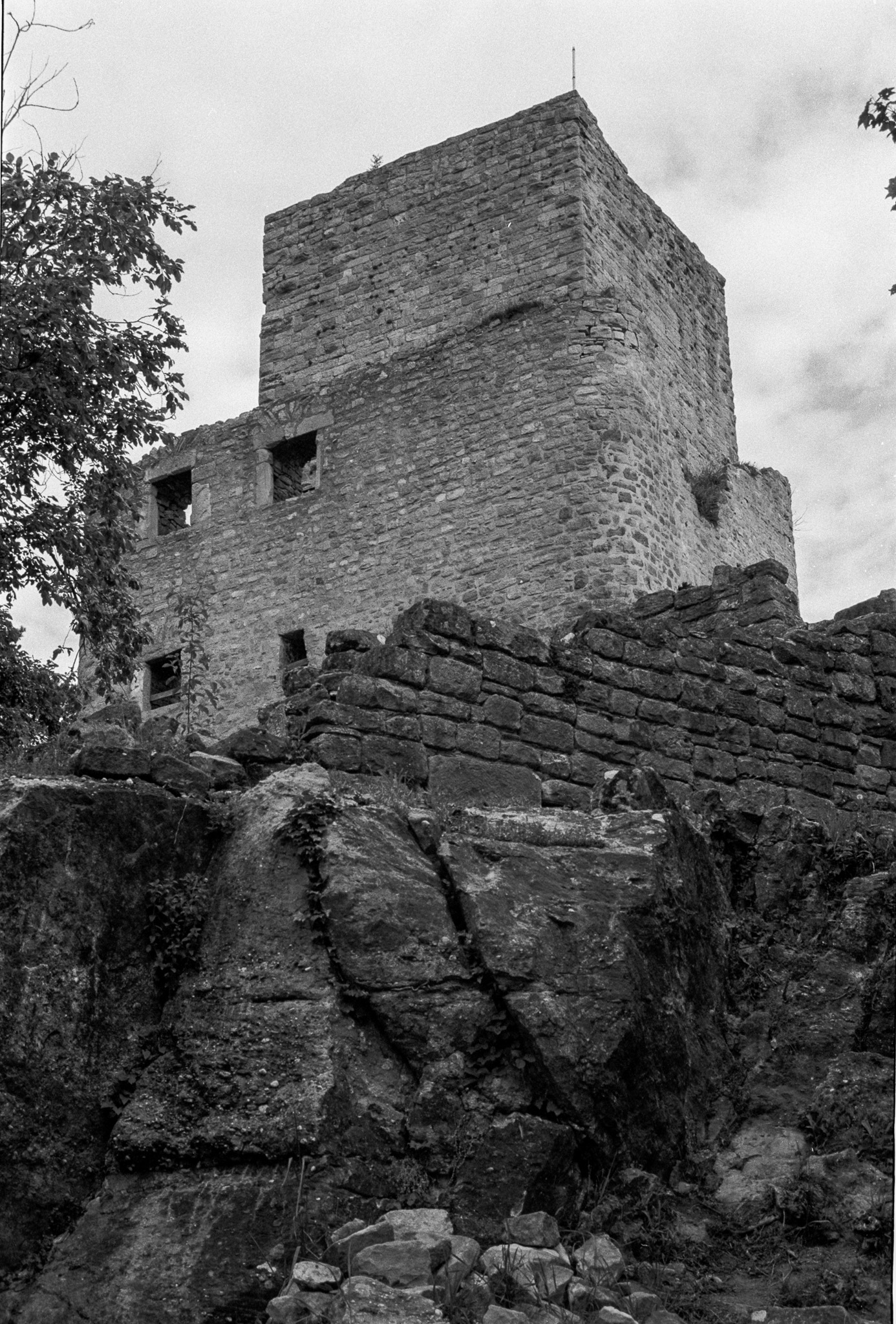 Tour massive d’une forteresse en ruine dominant un mur de pierre et un rocher en noir et blanc
