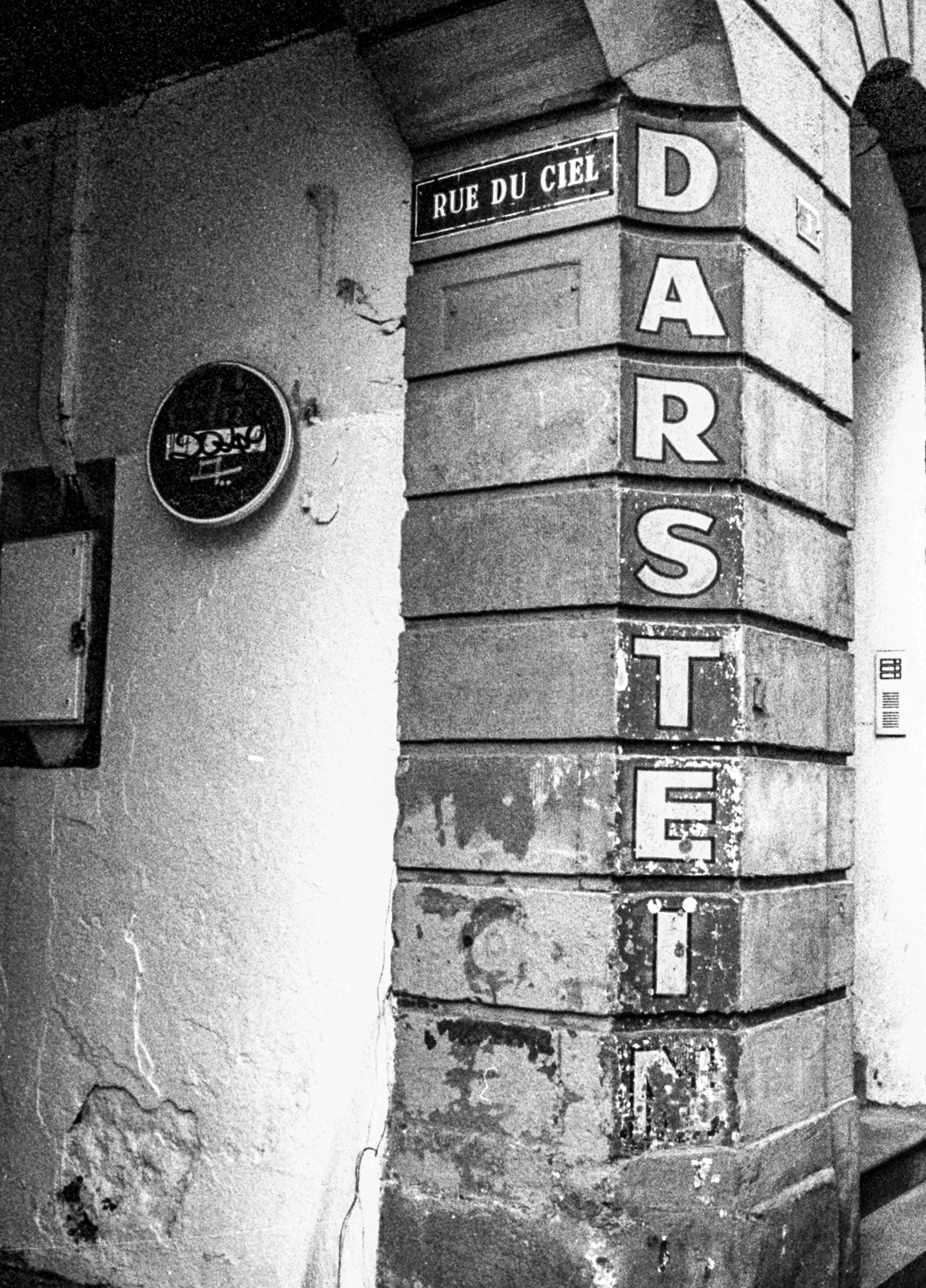 Plaque de la rue du Ciel et inscription verticale peinte sur l’angle d’un bâtiment photographié en noir et blanc