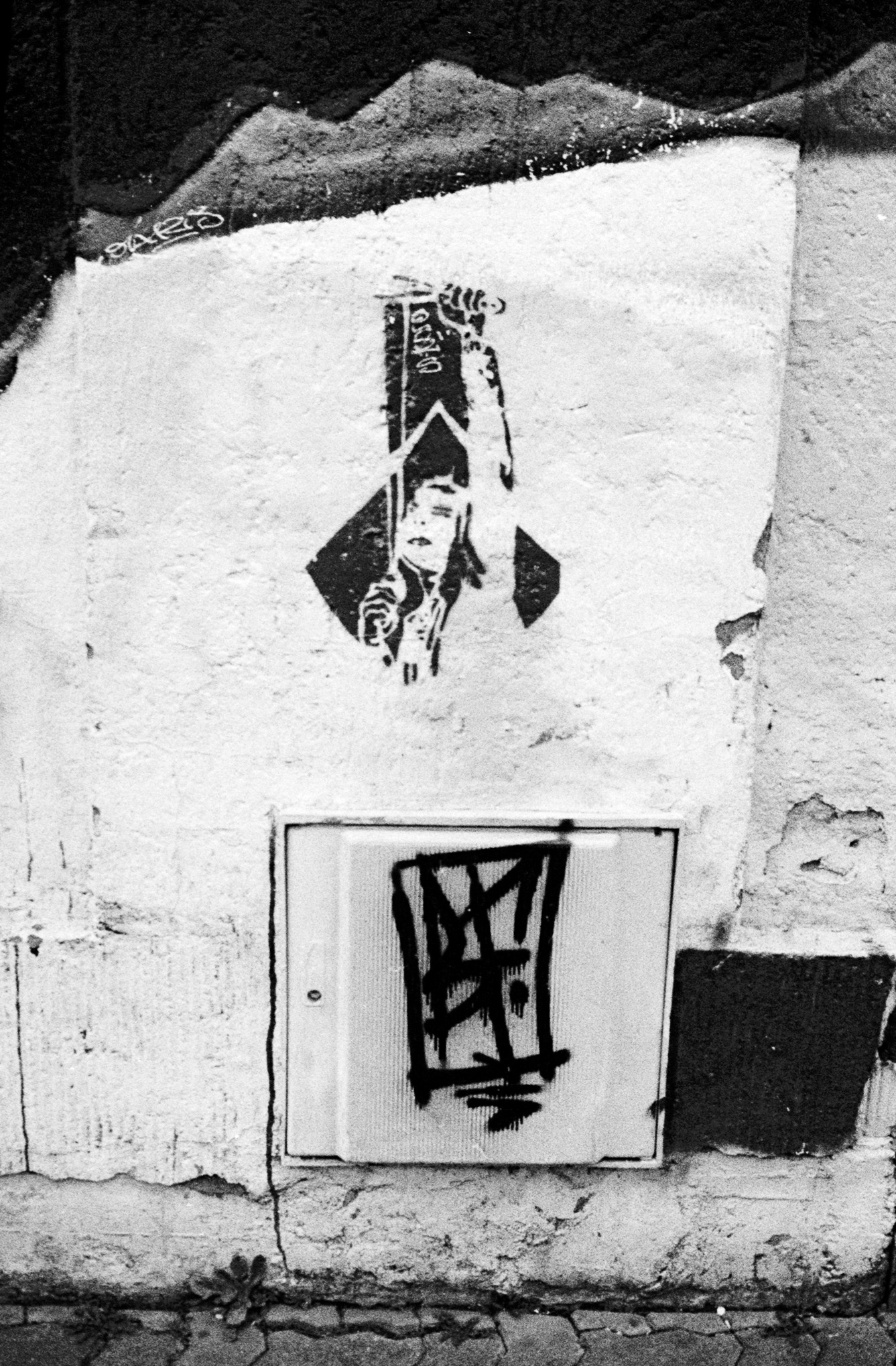 Pochoir noir représentant une silhouette sur un mur blanc, au-dessus d’un petit panneau couvert d’un graffiti