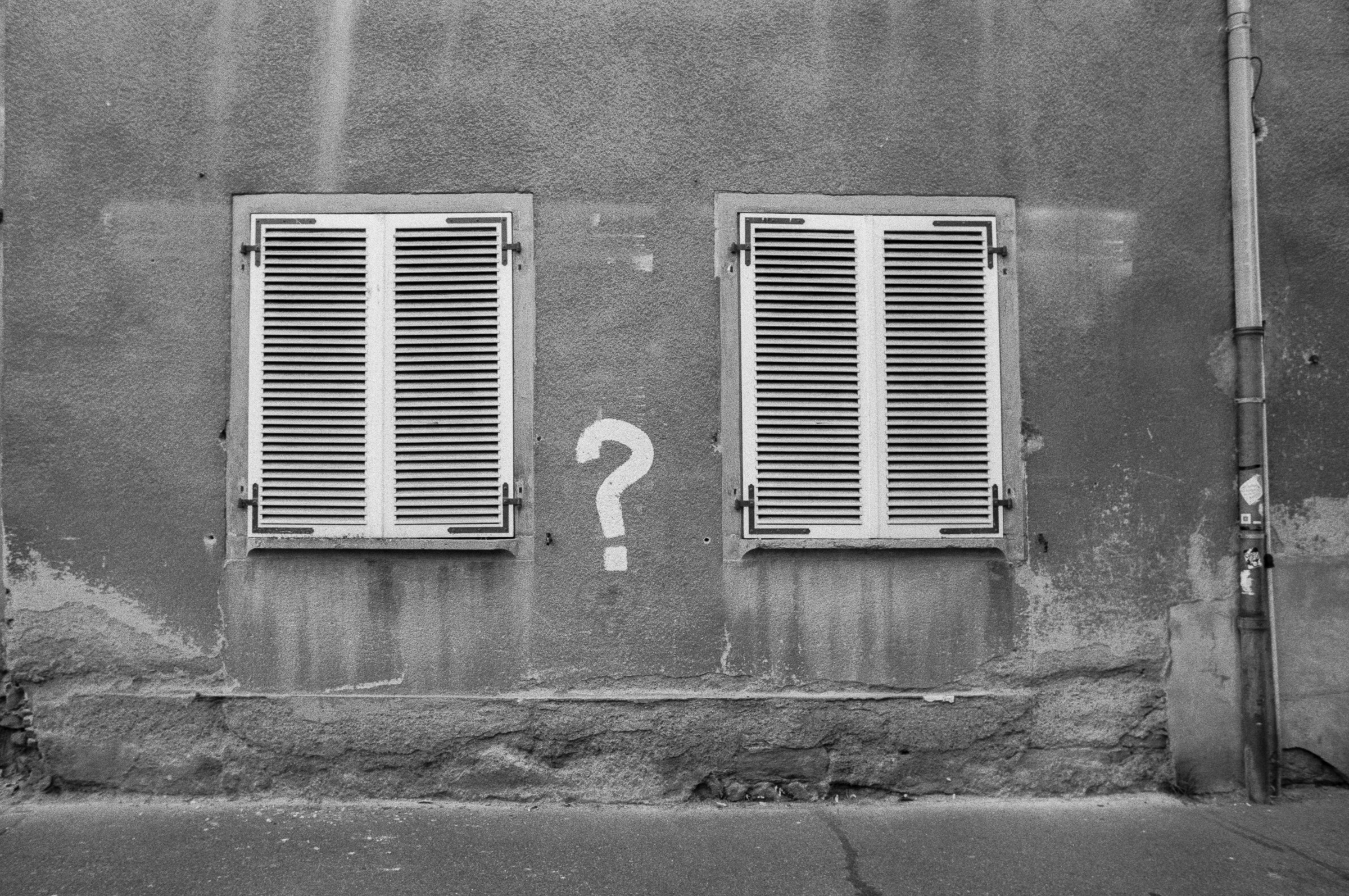 Deux fenêtres à volets fermés séparées par un point d’interrogation peint sur une façade ancienne